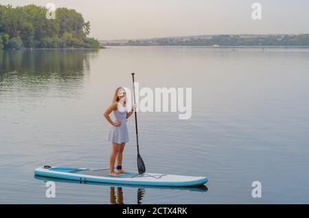 Young woman in a dress swims on the lake on surfboard Stock Photo