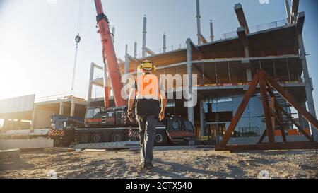 Back Shot of Worker Contractor Wearing Hard Hat and Safety Vests Walks on Industrial Building Construction Site. In the Background Crane, Skyscraper Stock Photo