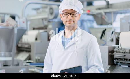 Young Male Quality Supervisor or Technician in Glasses is Working on a Food Factory. Employee Looks and Smiles at Camera. He Wears a White Sanitary Stock Photo