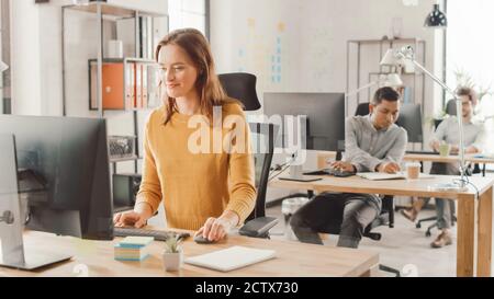 Beautiful and Smart Red Haired Female Specialist Sitting at Her Desk Works on a Desktop Computer. In the Background Modern Bright Office with Diverse Stock Photo