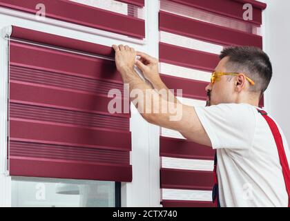 Young man in a uniform installing new window blinds. Professional service, home repair and renovation works Stock Photo