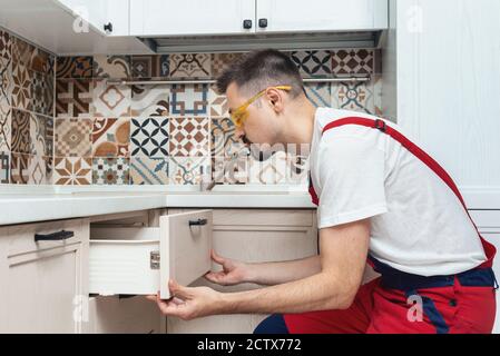 Worker dressed in workers' overall assembing furniture in kitchen. home and moving concept. installing new custom kitchen cabinet furniture decoration cabinets. Stock Photo