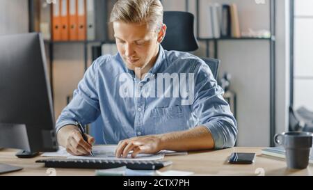 Handsome Blonde Businessman Sitting at His Desk in the Office Works on Desktop Computer, Working with Documents, Charts, Graphs, Statistic and Stock Photo