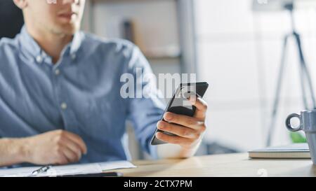 Entrepreneur Sitting at His Desk Works with Documents in the Stylish Office, Using Smartphone, Uses Social Media App, Emailing Business Partners Stock Photo