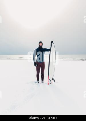 Winter and surfer with surfboard. Snowy beach with male surfer in wetsuit. Stock Photo