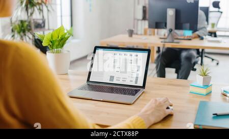 Over the Shoulder: Female Engineer Developer Sitting at Her Desk Using Laptop with Hardware Microchip Design. Office where Diverse Team of Young Stock Photo