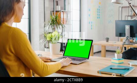 Over the Shoulder: Creative Young Woman Sitting at Her Desk Using Laptop with Mock-up Green Screen. Office where Diverse Team of Young Professionals Stock Photo