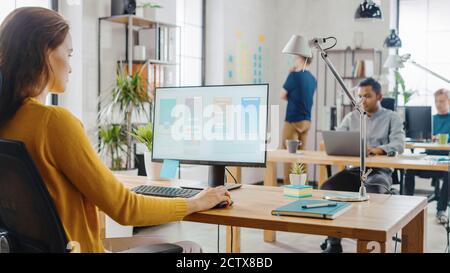 Over the Shoulder: Creative Young Woman Sitting at Her Desk Using Desktop Computer with Screen Showing Project Management, Statistics and Graphs Stock Photo