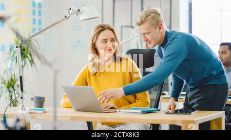 Beautiful and Creative Young Woman Sitting at Her Desk Using Laptop Computer, Young Handsome Team Leader Gives Advice about Project Details. Office Stock Photo
