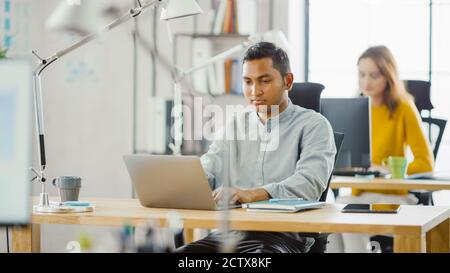 Smart and Handsome Indian Information Technology Specialist Sitting at His Desk works on a Laptop. In the Background Modern Office with Diverse Team Stock Photo