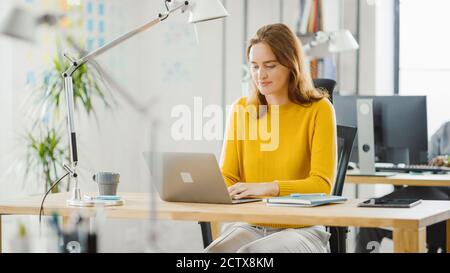Beautiful and Creative Young Woman Sitting at Her Desk Using Laptop Computer. In the Background Bright Office where Diverse Team of Young Stock Photo