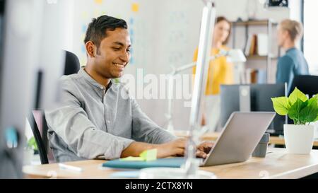 Handsome Smiling Indian Office Worker Sitting at His Desk works on a Laptop. In the Background Modern Office with Diverse Team of Young Professionals Stock Photo