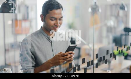 Portrait of Handsome Professional Indian Man Uses Mobile Phone, Writes Important Email. Successful Man Using Smartphone Working in Bright Diverse Stock Photo