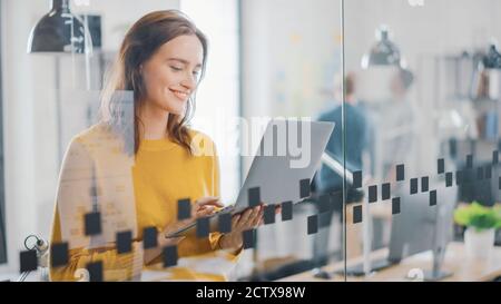 Portrait of Smiling Young Woman with Red Hair Wearing Yellow Sweater Uses Modern Laptop while Standing. Successful Woman Working in Bright Diverse Stock Photo