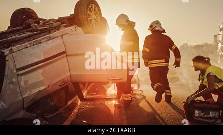 Car Crash Traffic Accident: Paramedics and Firefighters Plan Rescuing Passengers Trapped in Rollover Vehicle. Medics Prepare Stretchers and First Aid Stock Photo