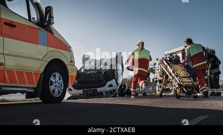 On the Car Crash Traffic Accident Scene: Team of Paramedics and Firefighters Rescue Injured People Trapped in Rollover Vehicle. Professionals Stock Photo