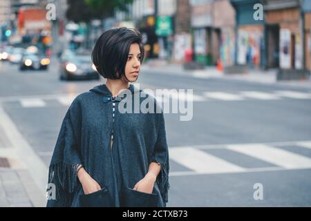 Beautiful Latin young woman in grey stylish poncho with fringe in urban street. Fashion and street style. Pretty hispanic brunette woman with short ha Stock Photo
