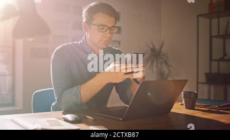 Professional Creative Man Sitting at His Desk in Home Office Studio Working on a Laptop. Man Using Smartphone to Check Social Media, Search Internet. Stock Photo