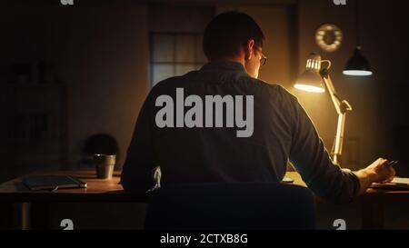 Back Shot of a Male Professional Designer Sitting at His Desk in Office Studio Working on a Laptop in the Evening. Man working with Data, Analyzing Stock Photo
