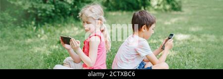 Boy and girl play games on smartphones outdoor. Kids digital gadget screen addiction. Children friends playing online in park. Contemporary problem Stock Photo