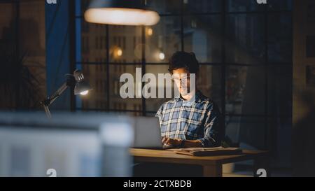 Handsome Male Creative Office Employee Sitting at His Desk Works on the Laptop. Evening in the Stylish Office Studio with Big Cityscape Window Stock Photo
