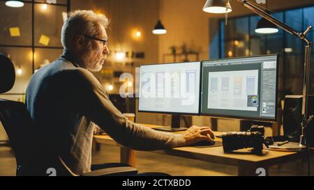 Creative Middle Aged Mobile Software Designer Sitting at His Desk Uses Desktop Computer with Two Screens Showing Smartphone Application Design Process Stock Photo