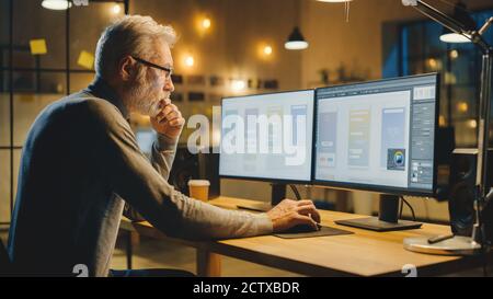 Creative Middle Aged Mobile Software Designer Sitting at His Desk Uses Desktop Computer with Two Screens Showing Smartphone Application Design Process Stock Photo