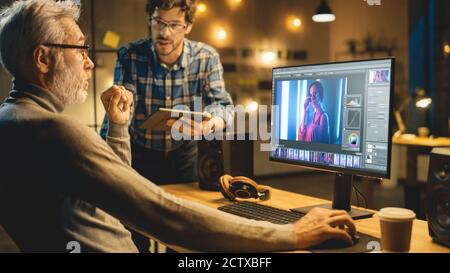 In the Evening Creative Middle Aged Photo Editor Works on a Desktop Computer Uses Image Editing Software, Has Conversation with Younger Specialist who Stock Photo