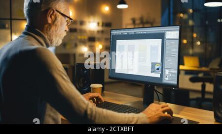 Creative Middle Aged Mobile Software Designer Sitting at His Desk Uses Desktop Computer with Screen Showing Smartphone Application Design Process Stock Photo