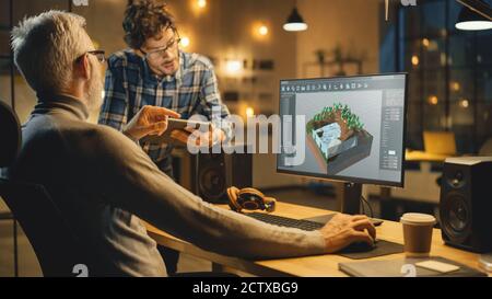 In the Evening Creative Middle Aged Video Game Developer Works on a Desktop Computer with Screen 3D Videogame Level Design, Has Conversation with Stock Photo