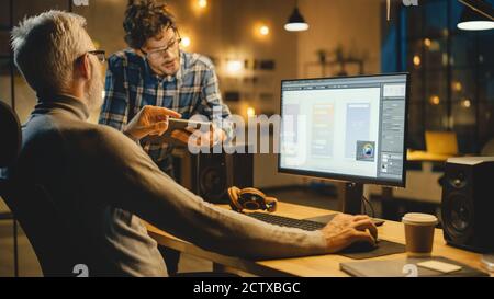 In the Evening Creative Middle Aged Mobile Software Designer Uses Desktop Computer with Smartphone Application Design, Has Conversation with Younger Stock Photo