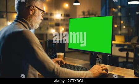 Creative Middle Aged Designer Sitting at His Desk Uses Desktop Computer with Green Mock-up Screen. In the Evening Creative Employe Working on Stock Photo