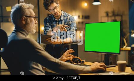 In the Evening Creative Middle Aged Man Works on a Desktop Computer Green Mock-up Screen, Has Conversation with Younger Specialist who Uses Tablet Stock Photo