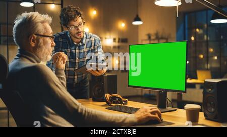 In the Evening Creative Middle Aged Man Works on a Desktop Computer Green Mock-up Screen, Has Conversation with Younger Specialist who Uses Tablet Stock Photo