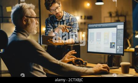 In the Evening Creative Middle Aged Mobile Software Designer Uses Desktop Computer with Smartphone Application Design, Has Conversation with Younger Stock Photo