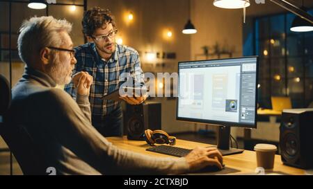 In the Evening Creative Middle Aged Mobile Software Designer Uses Desktop Computer with Smartphone Application Design, Has Conversation with Younger Stock Photo
