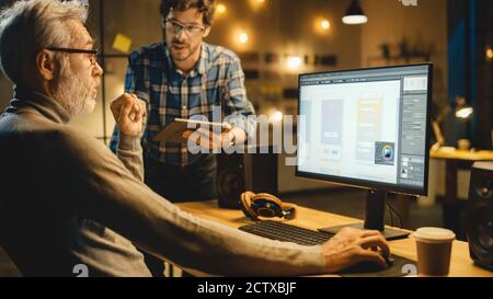 In the Evening Creative Middle Aged Mobile Software Designer Uses Desktop Computer with Smartphone Application Design, Has Conversation with Younger Stock Photo