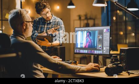 In the Evening Creative Middle Aged Photo Editor Works on a Desktop Computer Uses Image Editing Software, Has Conversation with Younger Specialist who Stock Photo
