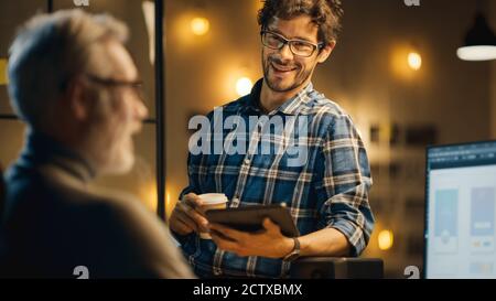 In the Evening Creative Middle Aged Smartphone Software Developer Works on a Desktop Computer with Screen Showing Application Design, Has Conversation Stock Photo