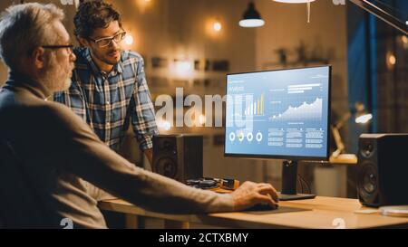 Professional Senior Business Martket Analyst Works on Desktop Computer Working with Charts, Graphs, Has Conversation with Younger Specialist who Gives Stock Photo