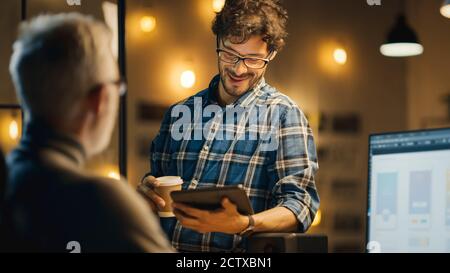 In the Evening Creative Middle Aged Mobile Software Designer Uses Desktop Computer with Smartphone Application Design, Has Conversation with Younger Stock Photo