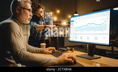 Professional Senior Business Martket Analyst Works on Desktop Computer Working with Charts, Graphs, Has Conversation with Younger Specialist who Gives Stock Photo