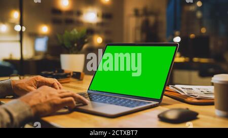 Close-up On the Hands of the Male Specialist Works on a Laptop Computer with Mock-up Green Screen. In the Background Evening in the Stylish Studio Stock Photo