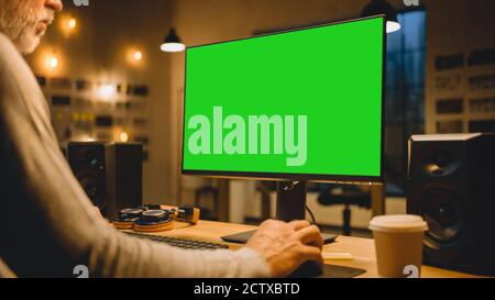 Creative Middle Aged Designer Sitting at His Desk Uses Desktop Computer with Two Green Mock-up Screens. Professional Office Employee Working Late in Stock Photo