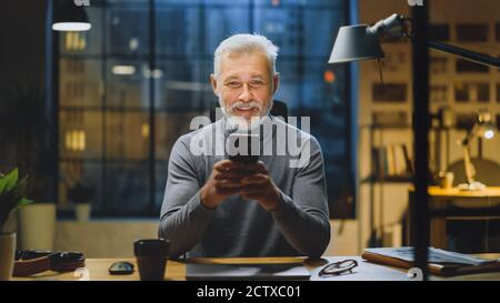 Portrait of the Handsome and Successful Middle Aged Bearded Businessman Uses Smartphone while Sitting at His Desk, Laughs and Smiles. Working from Stock Photo