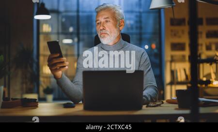 Portrait of the Handsome and Successful Middle Aged Bearded Businessman Working at His Desk Using Laptop Computer, Checks His Smartphone. Working from Stock Photo