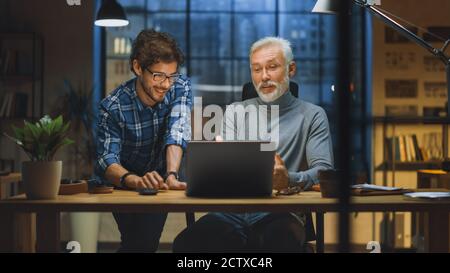 Handsome Middle Aged Entrepreneurand Successful Businessman Sitting at His Desk works on Laptop, Has Discussion with Younger Colleague. They Smile and Stock Photo