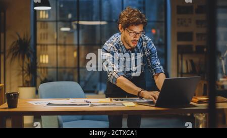 Young Creative Designer Working at His Desk, Focused on the Laptop and Dockuments, Choosing Right Design, Concept and Sketches. In the Background Stock Photo