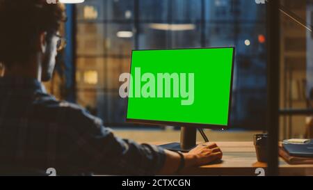 Over the Shoulder: Creative Young Man Sitting at His Desk Using Desktop Computer with Mock-up Green Screen. Evening in the Stylish Office Studio with Stock Photo