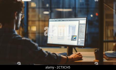Over the Shoulder Shot of a Young App Developer Using Desktop Computer, Creating New Mobile Phone Application Design, Software UI Development. Evening Stock Photo